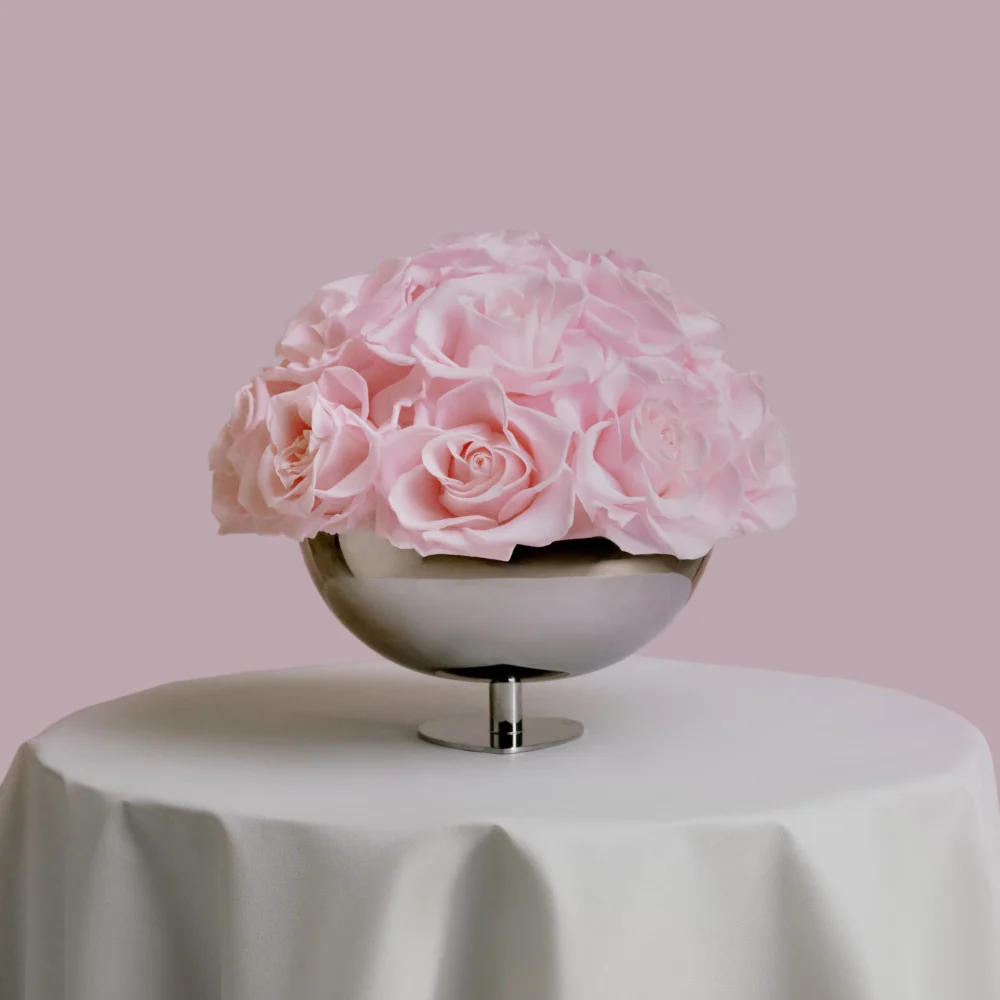 Metallic silver bowl filled with large realistic pink roses on white tablecloth-covered table with soft pink background