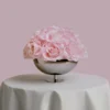 Metallic silver bowl filled with large realistic pink roses on white tablecloth-covered table with soft pink background
