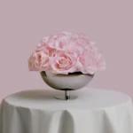 Metallic silver bowl filled with large realistic pink roses on white tablecloth-covered table with soft pink background