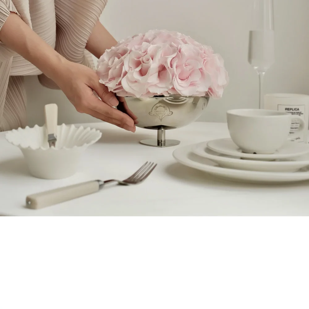 Woman in beige dress arranging pink hydrangeas in silver ornate vase on white table with champagne glass and plates