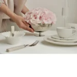 Woman in beige dress arranging pink hydrangeas in silver ornate vase on white table with champagne glass and plates