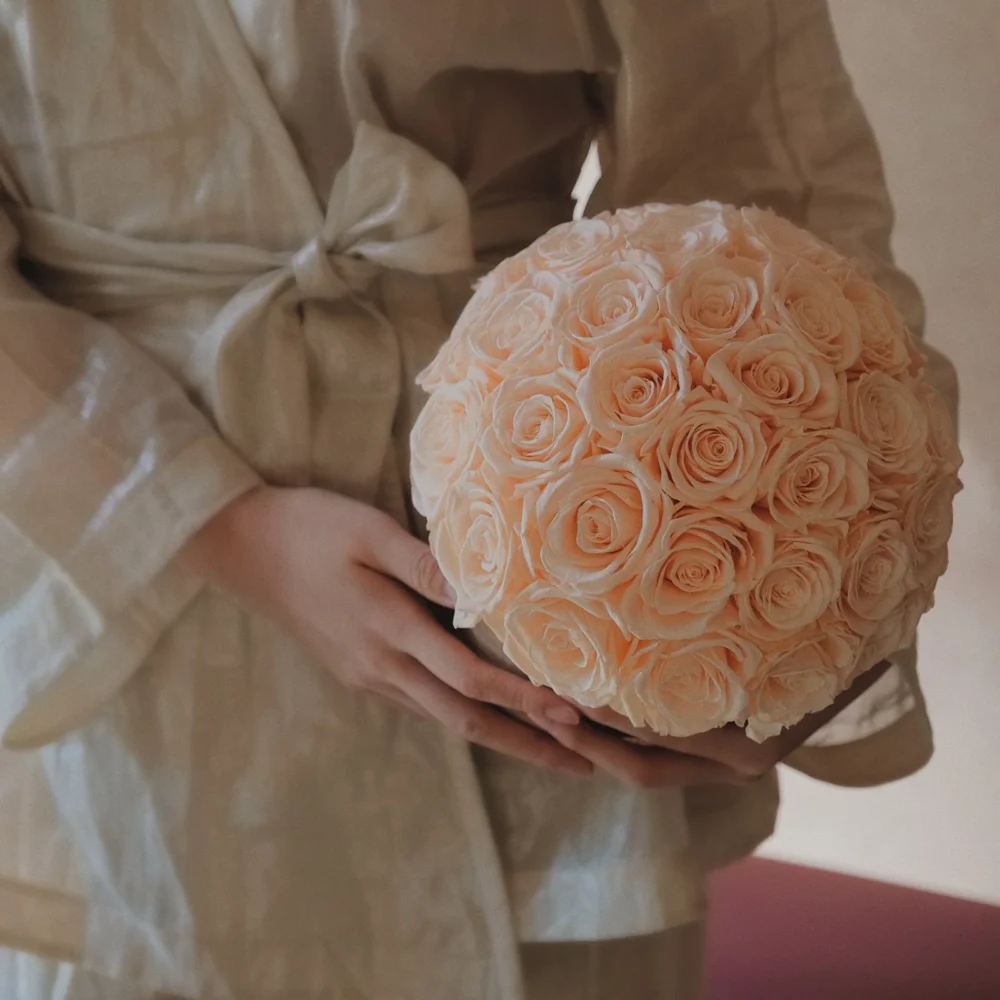Person in beige satin robe holding bouquet of soft pink roses against blurred background
