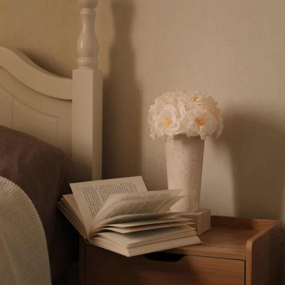 Serene bedroom with wooden nightstand holding open book and vase of delicate white flowers against cream wall