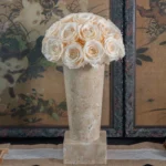 Tall cream ceramic vase with textured surface holding elegant white rose bouquet on dark wooden table with vintage Asian artwork