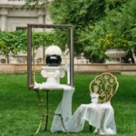 Whimsical outdoor setup with white vase in golden frame and vintage white chair. Lush green grass and stone fountain add a floral-themed serene ambiance.
