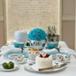 Beautifully arranged tea party setting with clear glass vase holding vibrant turquoise roses and silver utensils