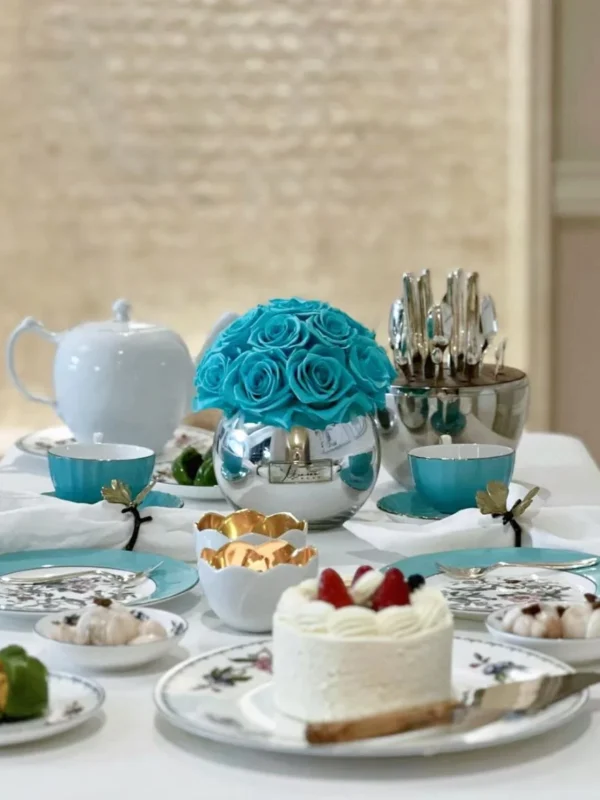 Beautifully arranged tea party setting with clear glass vase holding vibrant turquoise roses and silver utensils