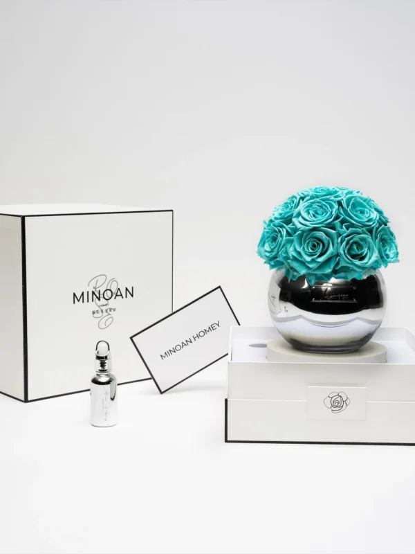 Minimalist elegant setup with clear glass vase of turquoise artificial roses beside "Minoa by Maison Margiela" box