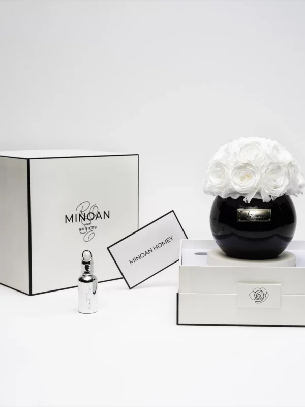 Photograph of a minimalist, modern interior setting featuring a black, glossy vase with a bouquet of white roses, positioned on a white, rectangular tray. To the left, a sleek, white box with a black trim labeled "Minoan" and "Minoan Home." In front of the box, a silver perfume bottle and a small, white, rectangular card with "Minoan Home" in black text. The background is plain white, emphasizing the elegant, sophisticated design.high quality, detailed, photograph , hd, 8k , 4k , sharp, highly detailed