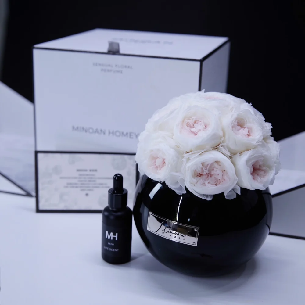Sleek black vase filled with white roses beside small black "MH" labeled bottle on dark background