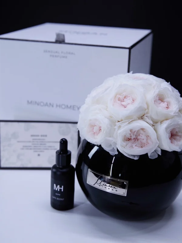 Sleek black vase filled with white roses beside small black "MH" labeled bottle on dark background