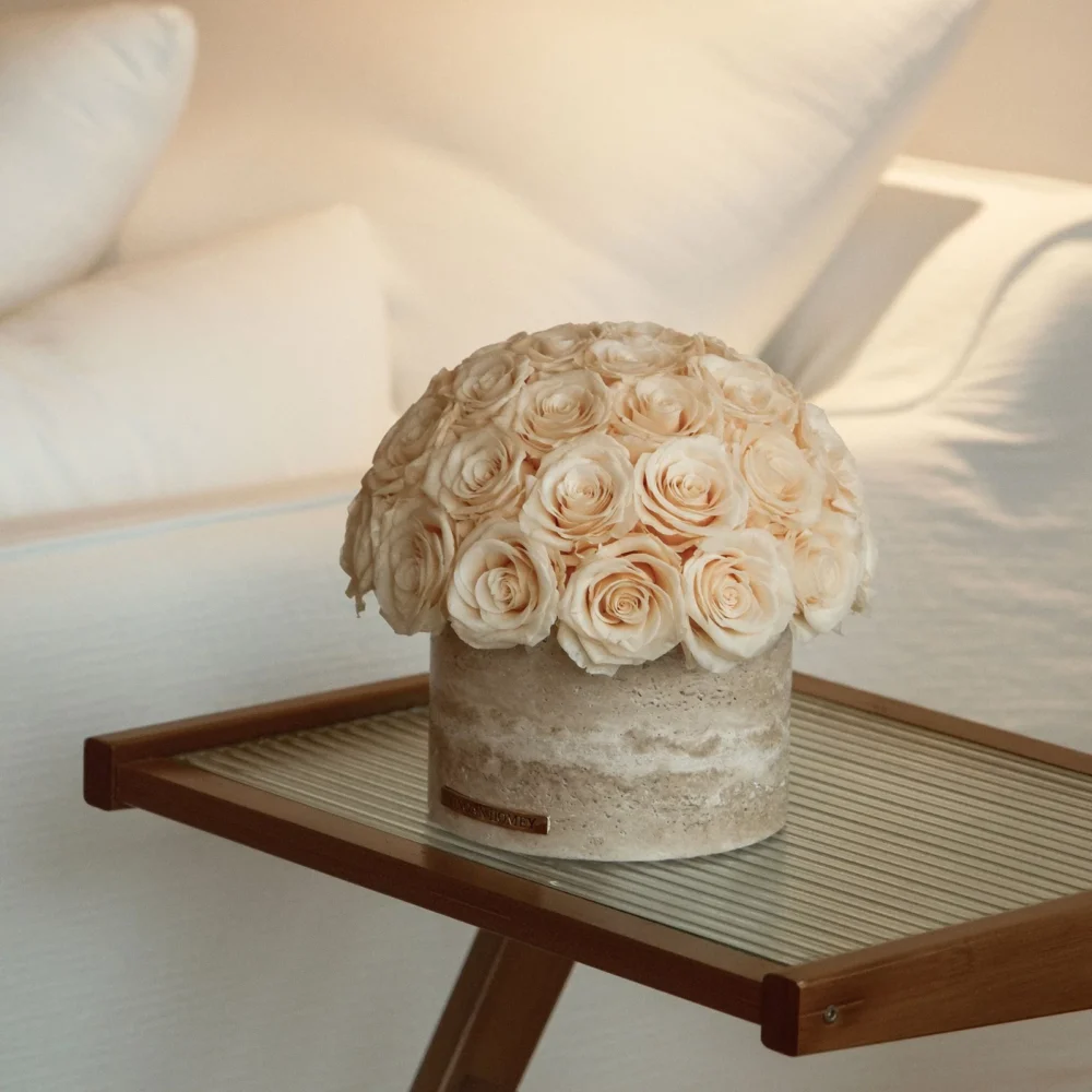 Minimalist bedroom corner with round wooden table holding cream rose bouquet in textured beige vase
