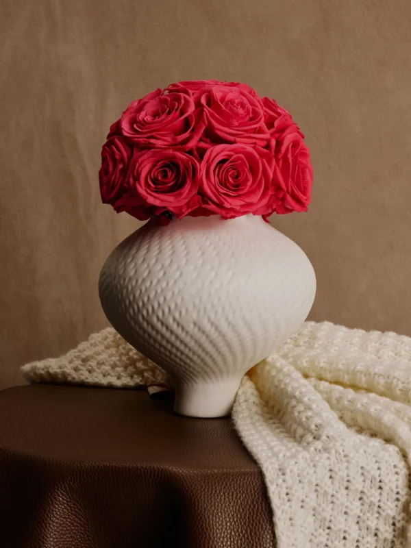White ceramic textured vase with vibrant red roses on brown leather tablecloth creating rustic atmosphere
