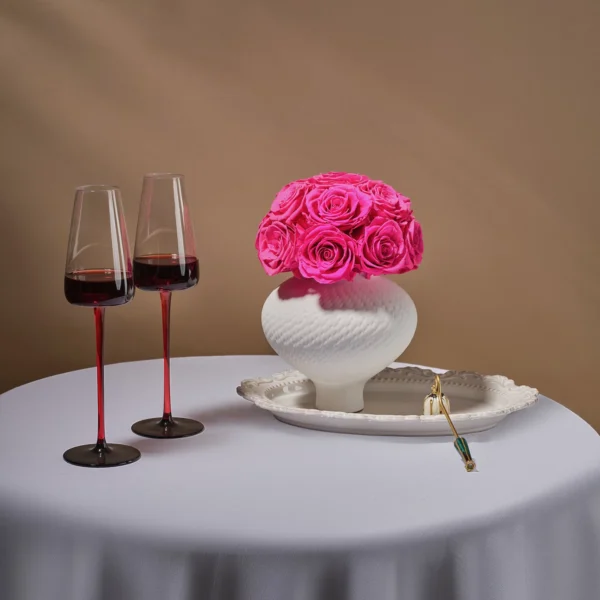 White ceramic vase with pink roses beside ornate plate with gold spoon and tulip wine glasses