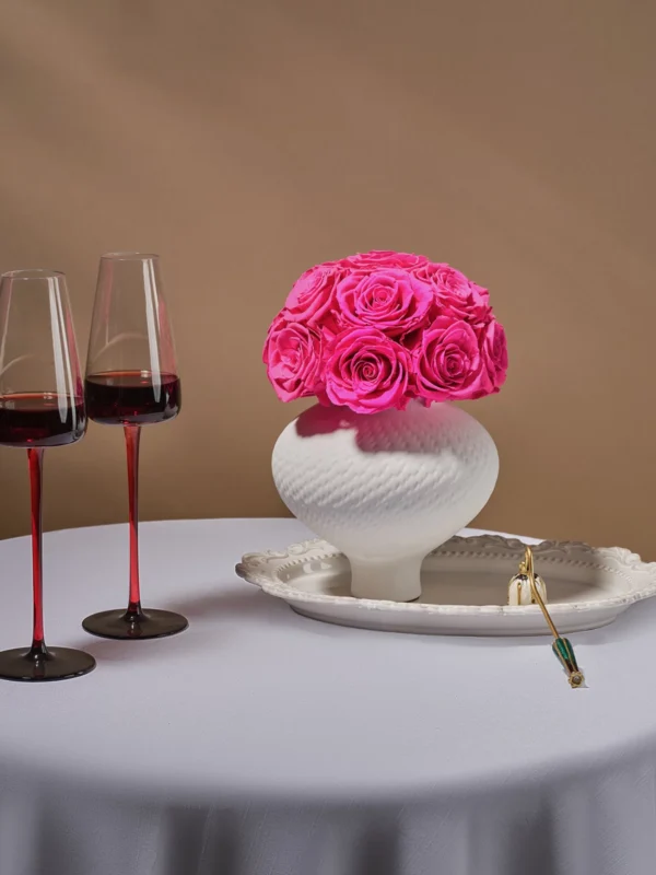 White ceramic vase with pink roses beside ornate plate with gold spoon and tulip wine glasses