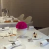 This photograph showcases an elegantly set dining table in a modern, minimalist restaurant. The table is draped in a white tablecloth, adorned with a vibrant, pink rose in a white ceramic vase.