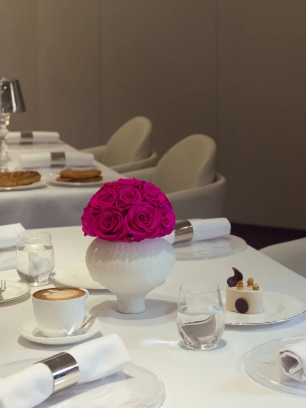 This photograph showcases an elegantly set dining table in a modern, minimalist restaurant. The table is draped in a white tablecloth, adorned with a vibrant, pink rose in a white ceramic vase.