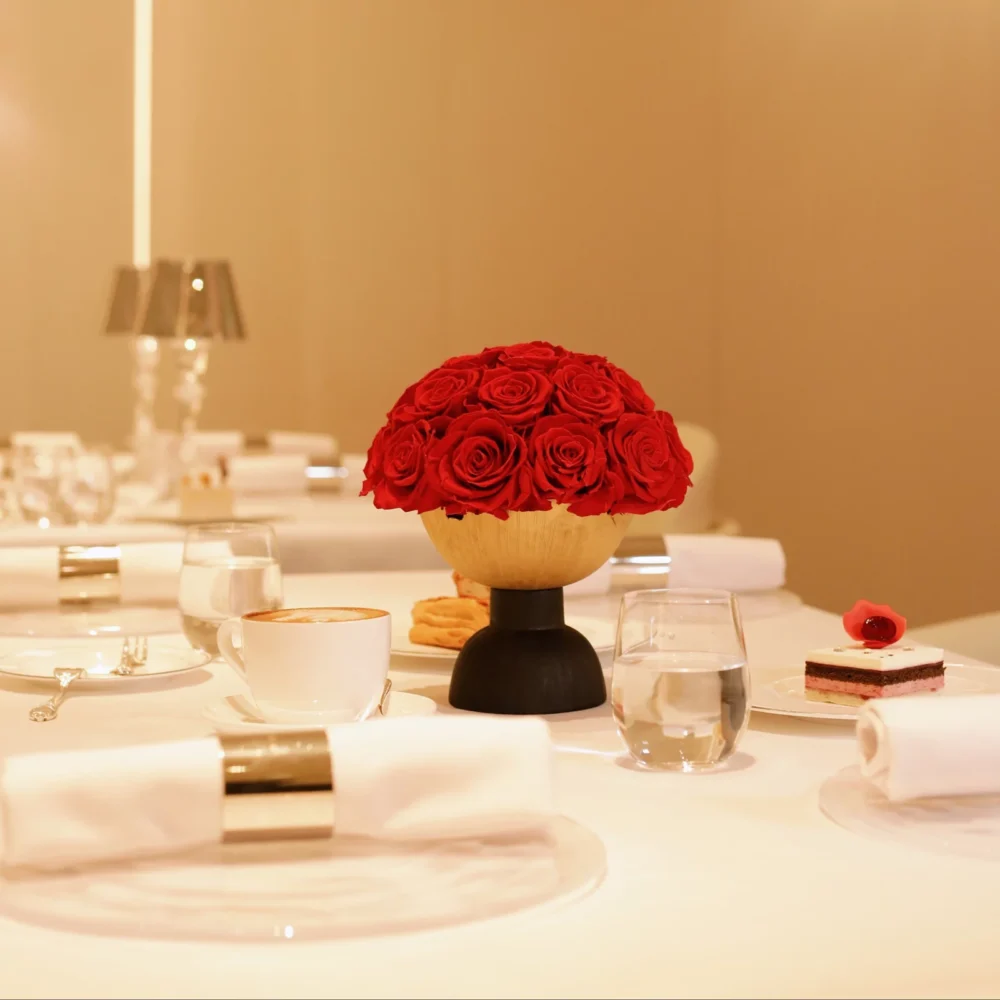 This photograph showcases an elegant table setting in a warm, beige-toned room. The centerpiece is a vibrant bouquet of red roses in a modern, wooden bowl.