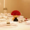 This photograph showcases an elegant table setting in a warm, beige-toned room. The centerpiece is a vibrant bouquet of red roses in a modern, wooden bowl.