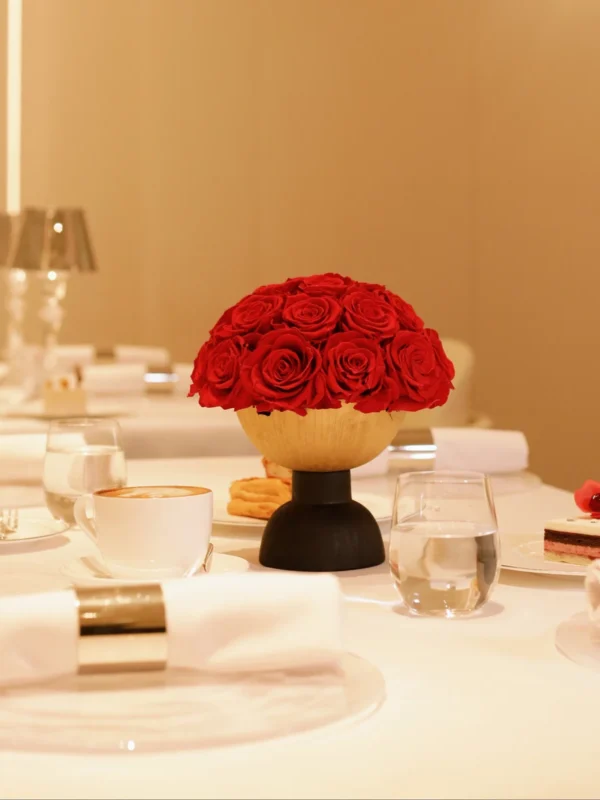 This photograph showcases an elegant table setting in a warm, beige-toned room. The centerpiece is a vibrant bouquet of red roses in a modern, wooden bowl.