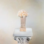 Pale peach vase with cream roses on white marble column. Classical floral designs and beige wall create a floral-themed sophisticated display.