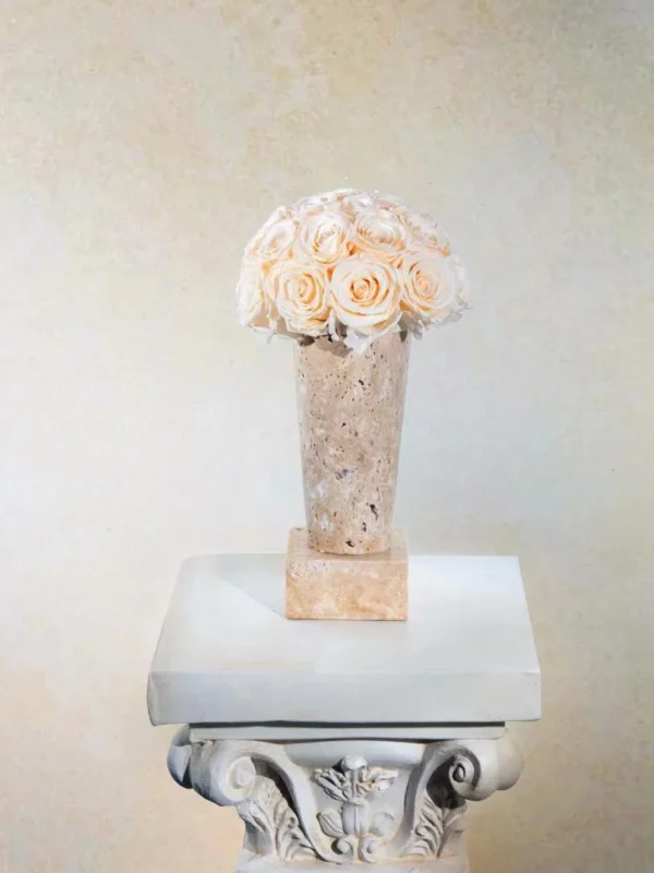 Pale peach vase with cream roses on white marble column. Classical floral designs and beige wall create a floral-themed sophisticated display.
