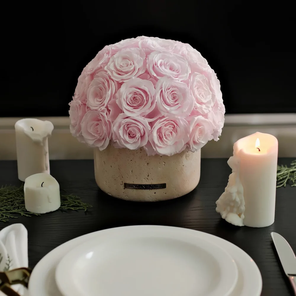 Delicate pink rose arrangement in rustic beige ceramic pot with white pillar candles on dark table