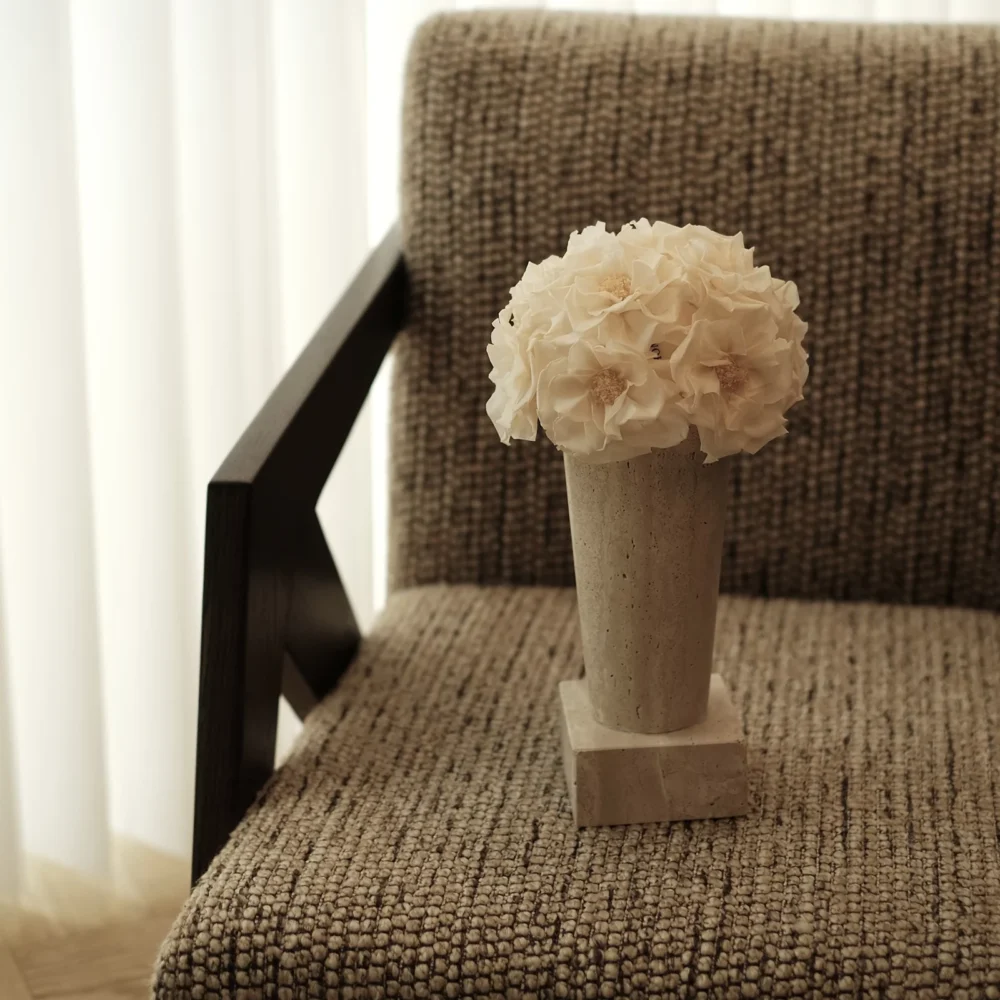 Minimalist beige fabric chair with black wooden armrest holding small ceramic vase of cream hydrangeas, soft natural light from white blinds