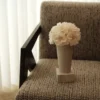 Minimalist beige fabric chair with black wooden armrest holding small ceramic vase of cream hydrangeas, soft natural light from white blinds