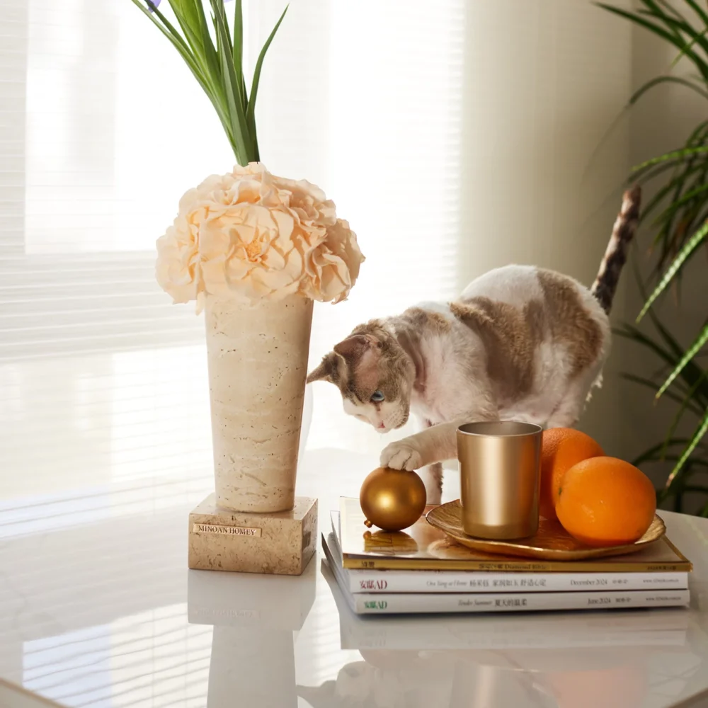 Grey and white cat on glass table next to beige vase with cream flowers and gold candle