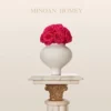 A minimalist white fluted vase with pink roses on a gold-accented marble pedestal, against a cream background. Elegant 'MINOAN HOMEY' text above. "