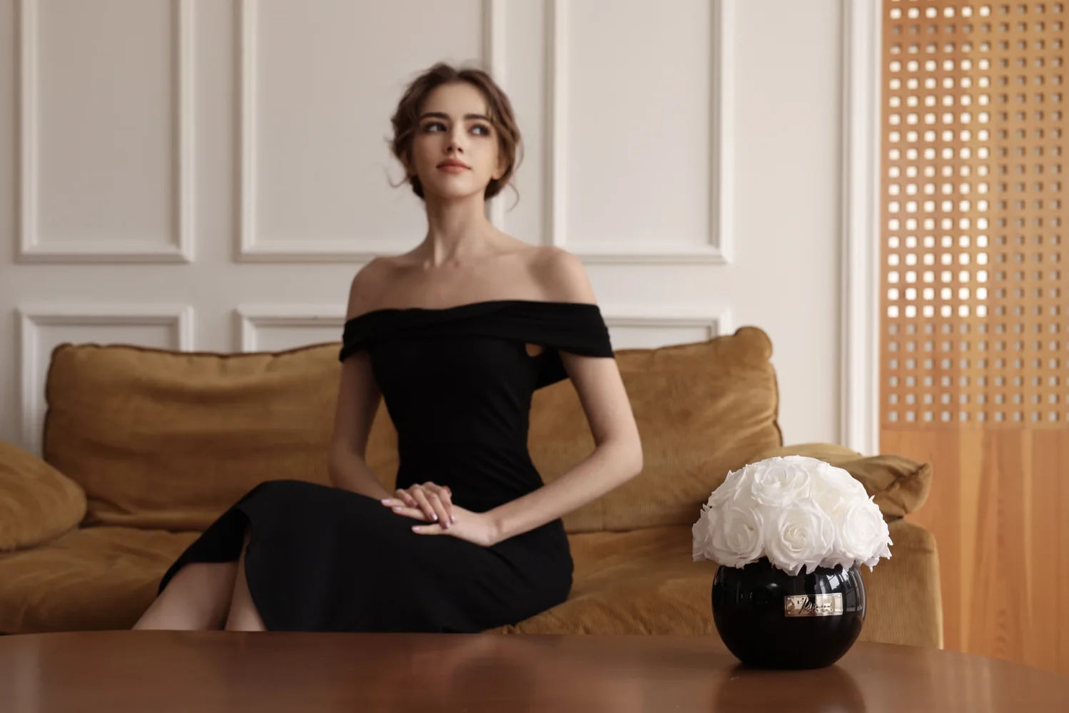 Slender woman in black dress sitting on mustard-yellow sofa holding black vase with white roses
