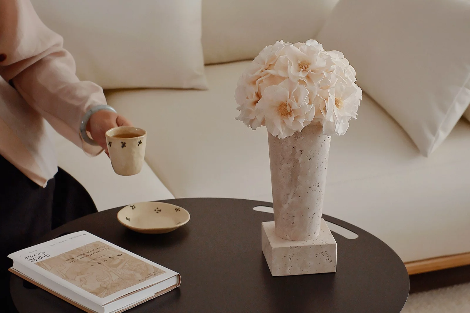 Person holding ceramic cup beside round black table with vase of white flowers