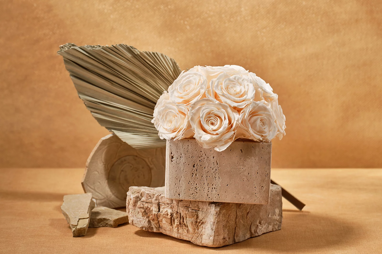 Minimalist earthy aesthetic with cream artificial rose bouquet on rectangular stone block surrounded by dried palm fronds