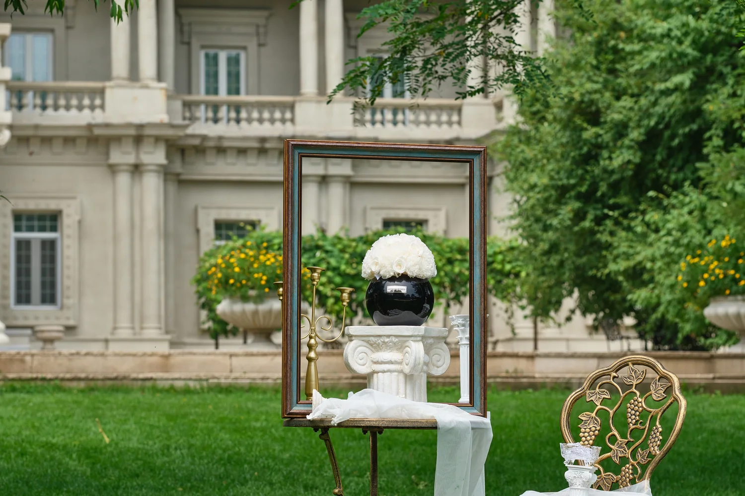 Vintage-style table with gold frame and black vase of white flowers. Classical mansion and lush garden enhance the floral-themed refined setting.