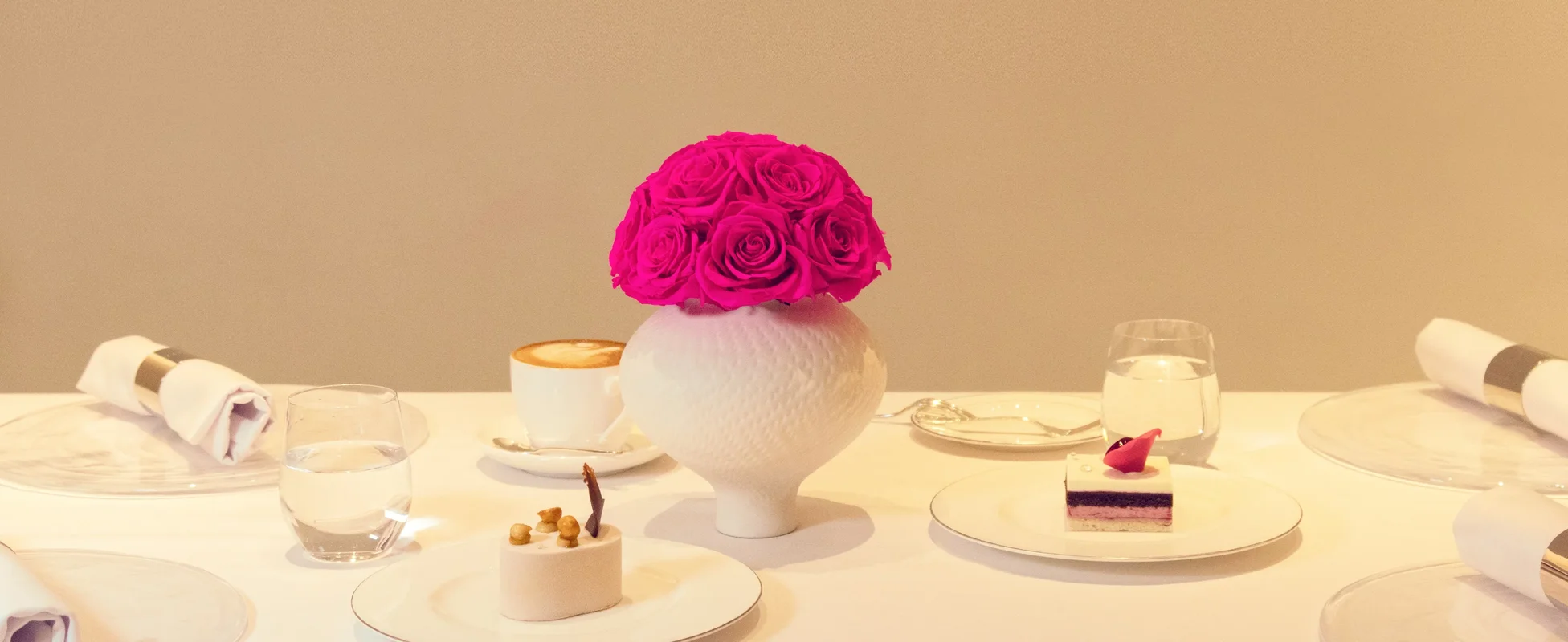 White ceramic vase with single pink rose beside dessert plates in minimalist setting