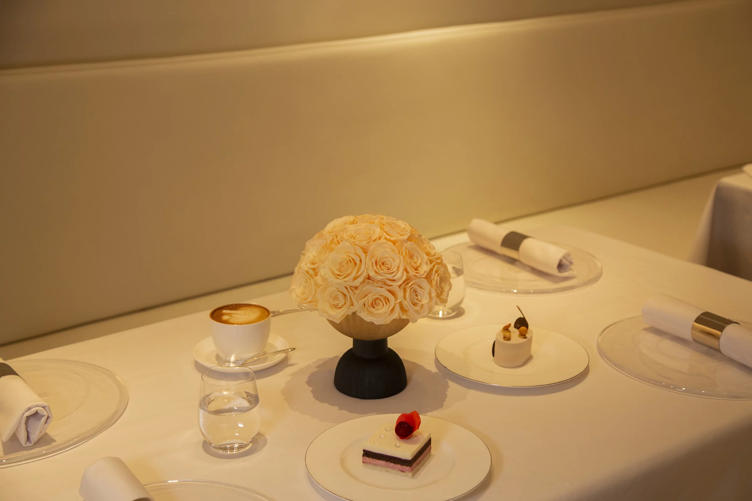 A photograph captures a minimalist, elegant dining table set for two. The table is draped in a white cloth, featuring a centerpiece of white roses in a black vase.