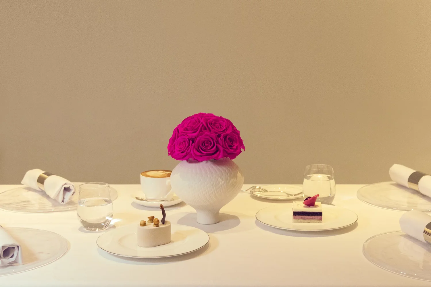 This photograph captures a sophisticated dining table setting, featuring a white ceramic vase filled with vibrant pink roses.
