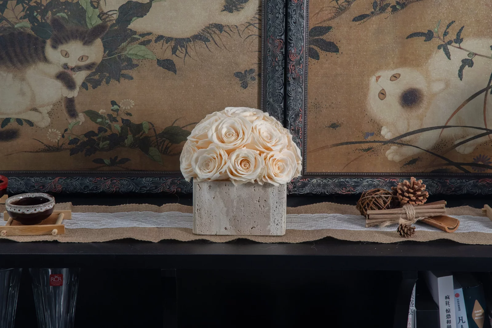 Rustic setup with white ceramic vase of cream fake roses on black shelf with pine cones and vintage Asian art backdrop