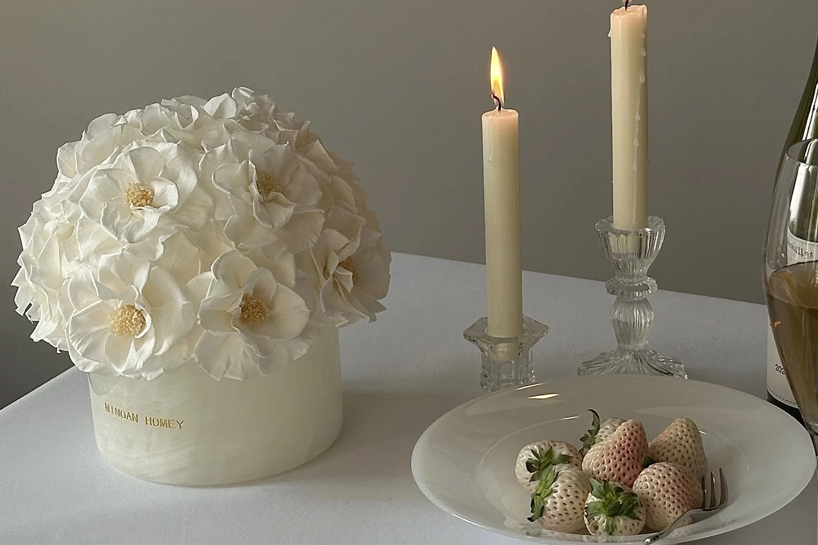 Sophisticated dining setup with tall white candles in glass holders flanking bouquet of white flowers in vase, champagne bottle and strawberries